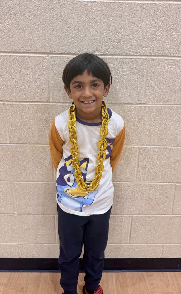 A child stands indoors, smiling, with a gold chain around their neck, wearing a white and orange t-shirt.