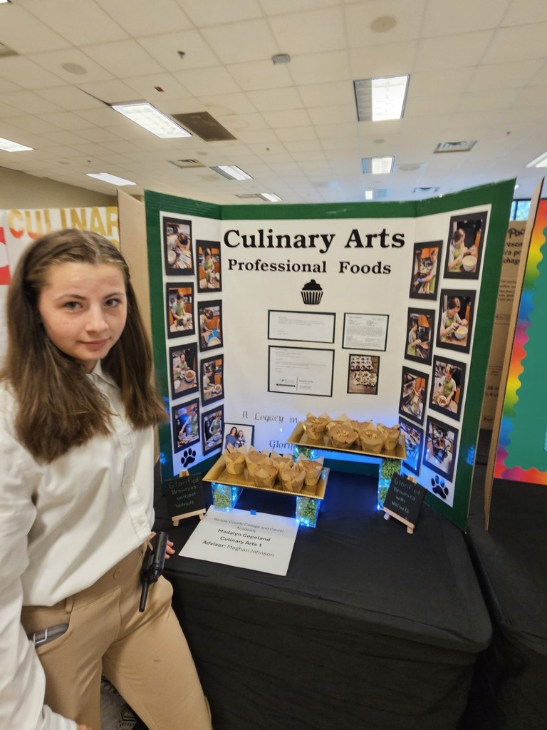 A person stands next to a display board titled "Culinary Arts Professional Foods". The board features pictures and text.