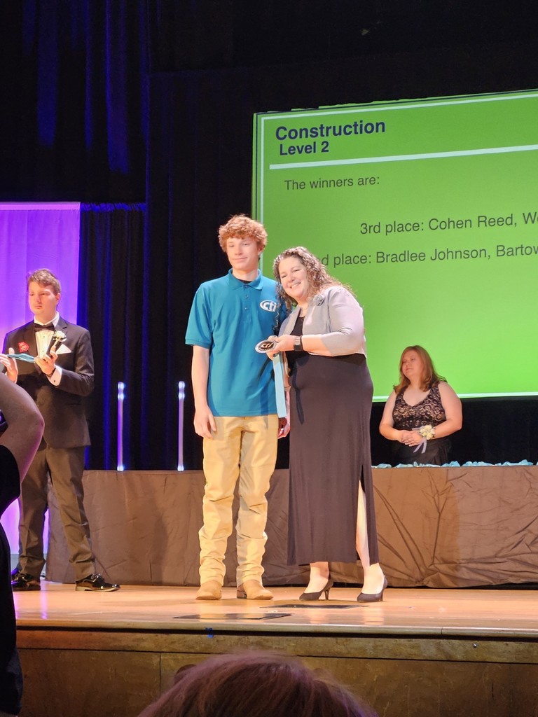 Three people stand on stage with a green screen behind them. One holds a trophy, and two others watch.
