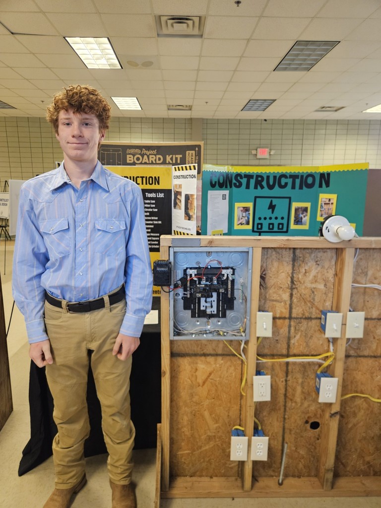 A young man stands in a classroom next to a display of electrical wiring and outlets.