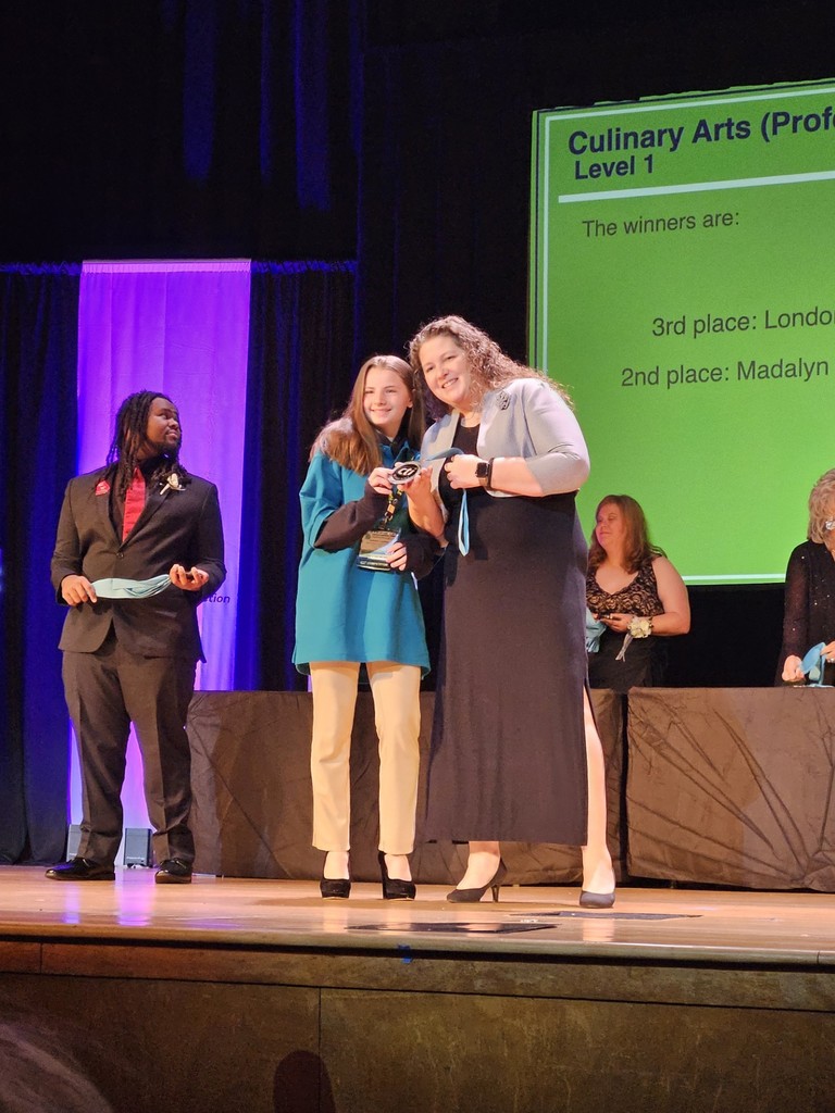 Winners of a culinary arts competition on stage. One woman holds a plaque. Behind them is a screen with winners' names.