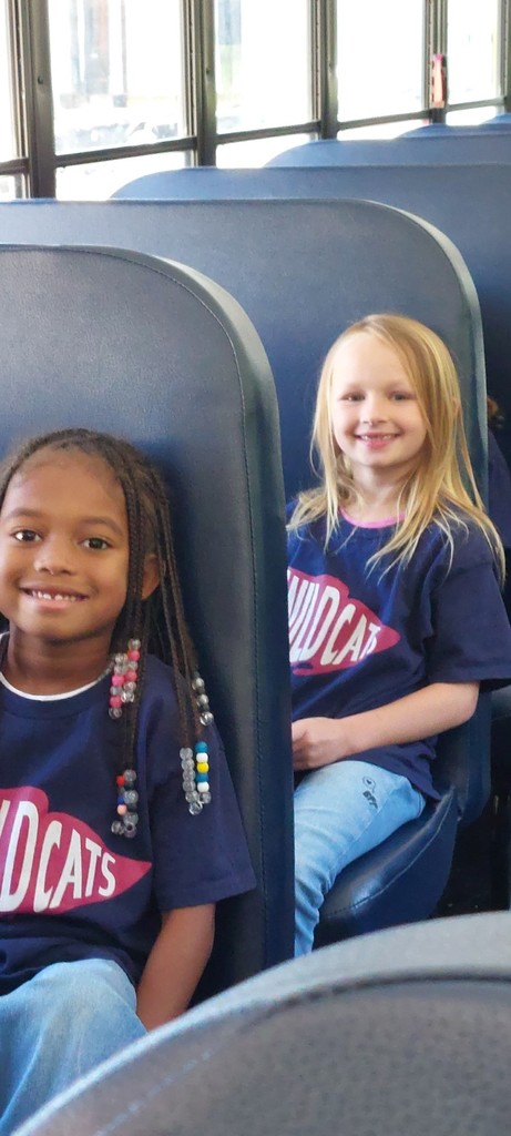 Two girls in blue Wildcats shirts sit in seats on a bus. One wears a necklace with colorful beads.