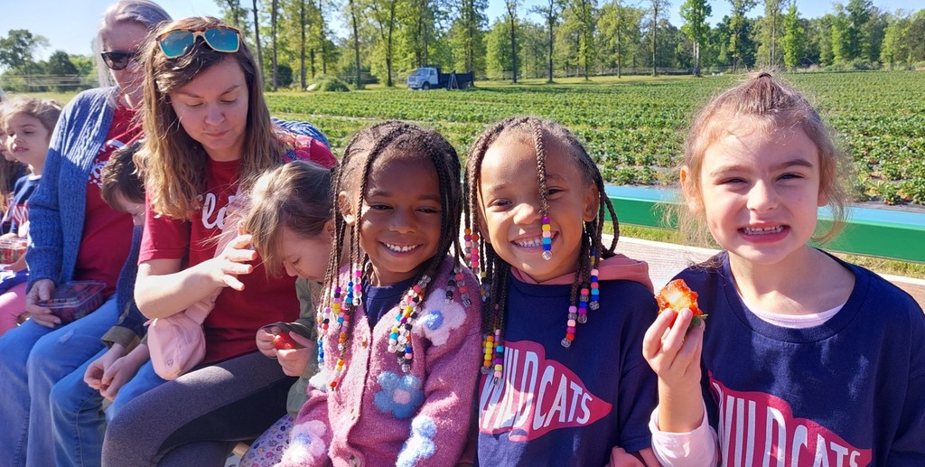 Five children and an adult woman smile and hold food outside. They sit on a bench. Trees and a field are in the background.