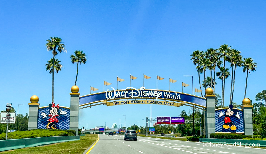 Entrance to Walt Disney World, featuring an archway with Mickey Mouse and Minnie Mouse designs, palm trees, and a clear blue sky.