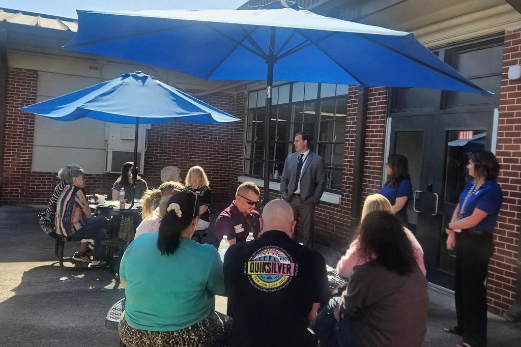 Group of people seated under blue umbrellas in front of a brick building, with a person standing nearby.