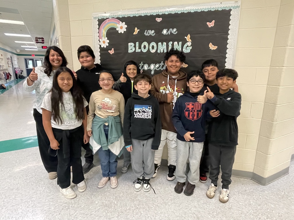 A group of children and adults pose for a photo in a hallway with a decorative sign reading "We are blooming."