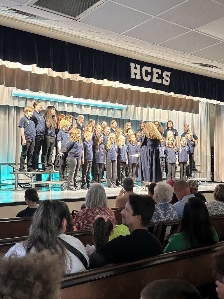 A choir on stage singing, with people seated in the audience. The stage has a curtain backdrop with "HCES" written above.