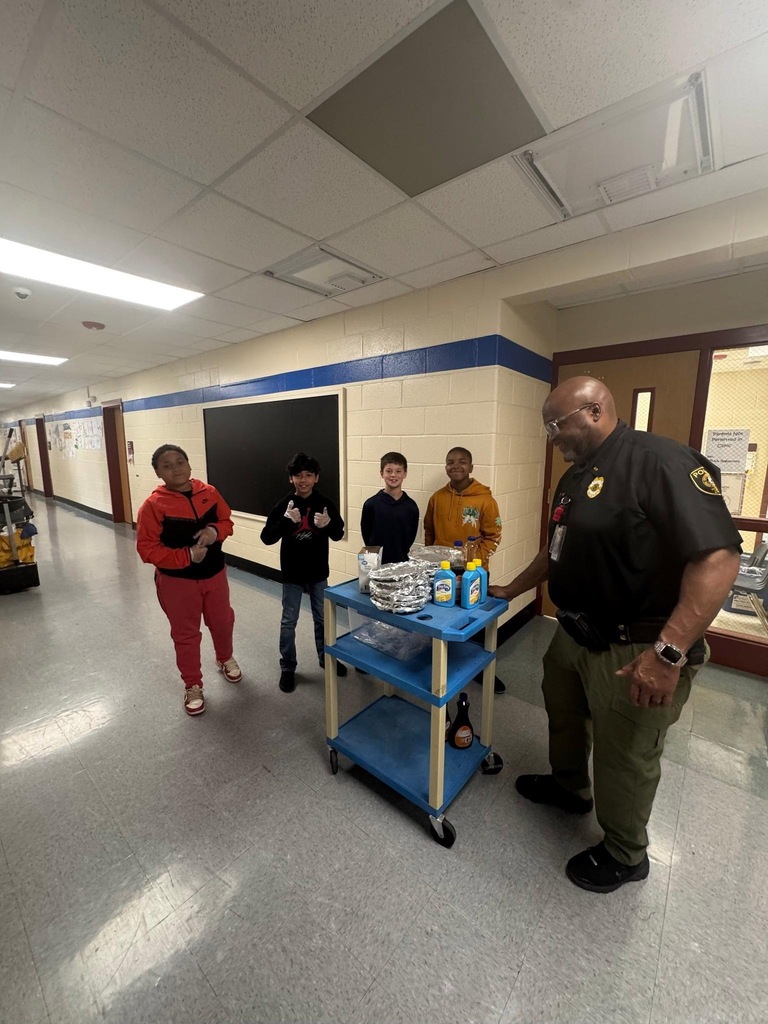 A man stands behind a blue cart with cleaning supplies, interacting with students in a hallway.