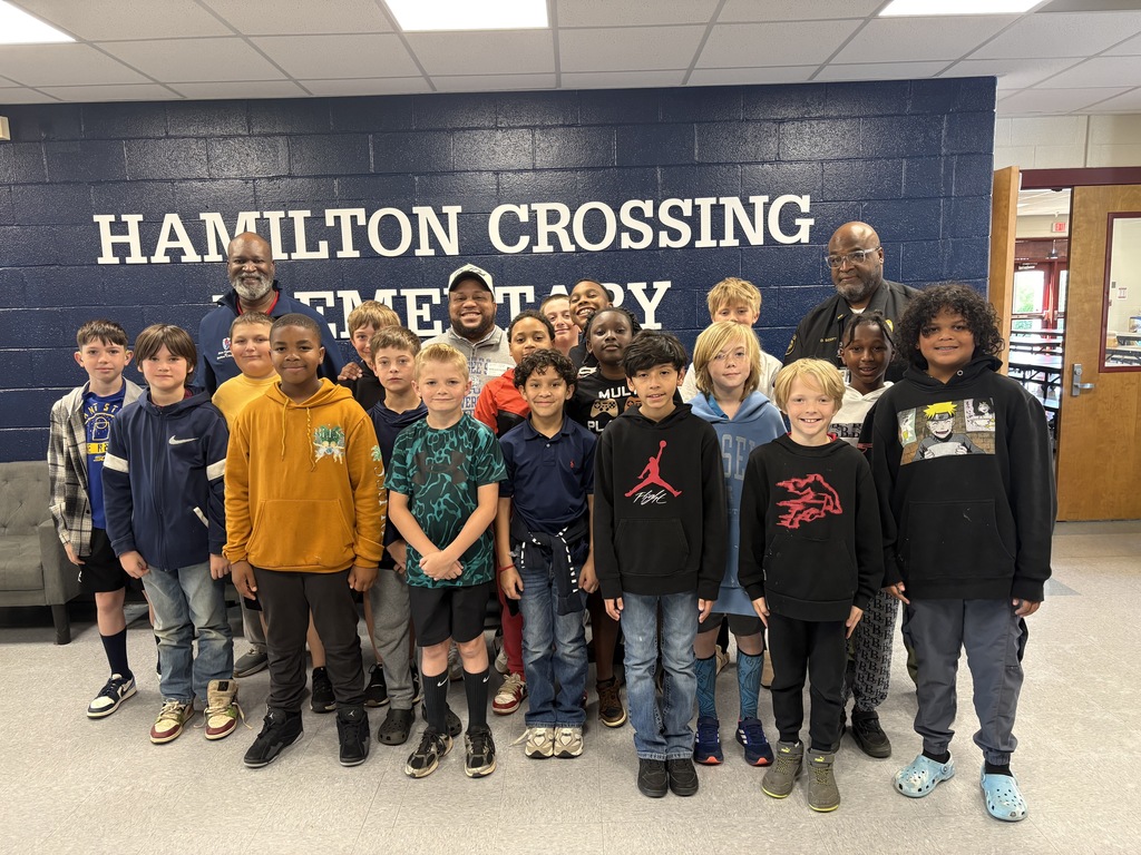 A group of children stands in a room with a blue wall reading "Hamilton Crossing". They wear casual clothing.