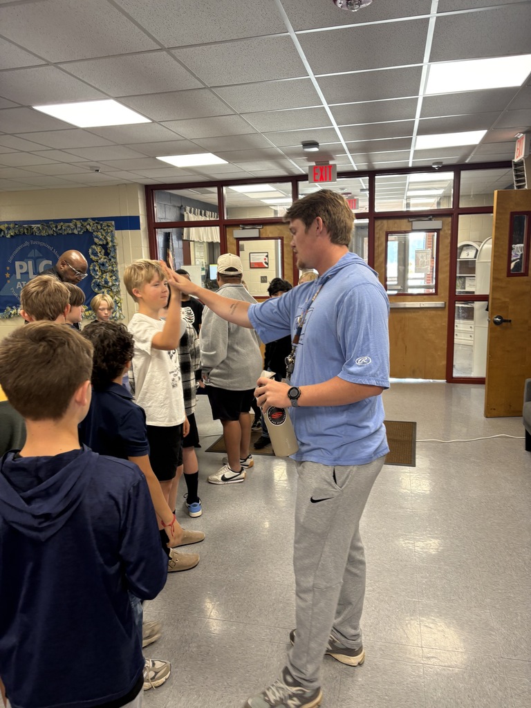 A man stands in a room, gesturing while addressing a group of children. The setting has white walls and tiled floors.