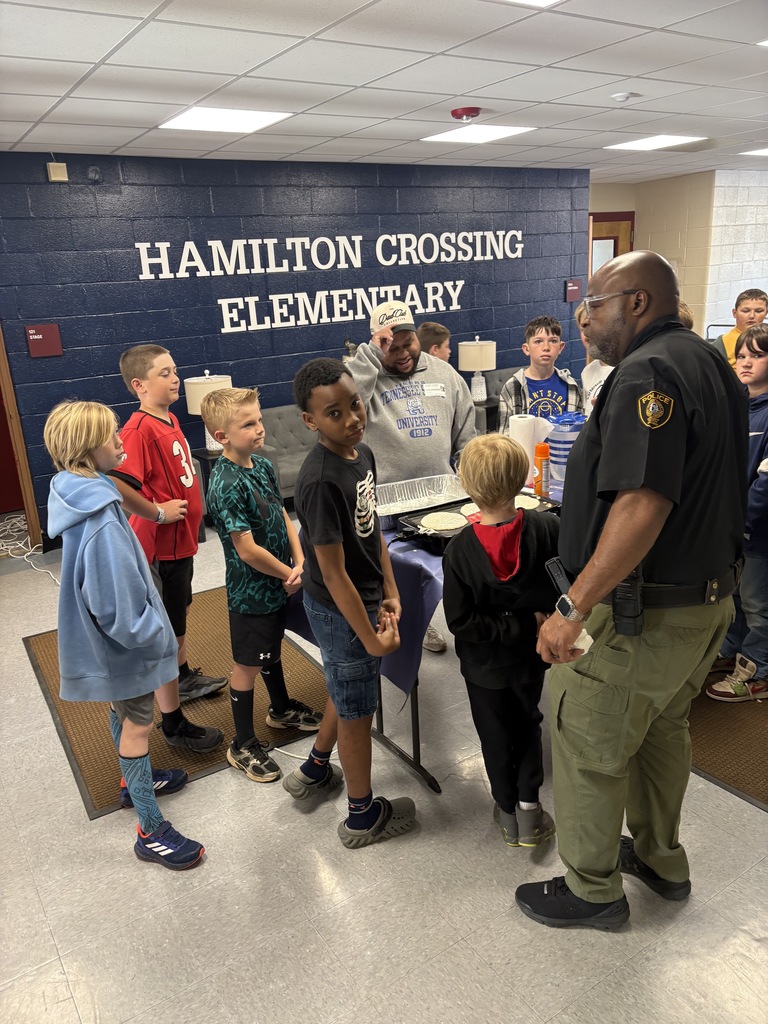 A police officer stands with a group of children at an event, with a banner behind reading "HAMILTON CROSSING ELEMENTARY."