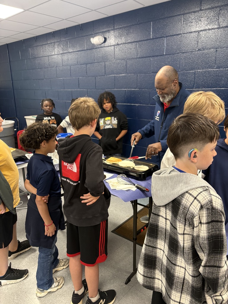 People gathered around a table with food, an older man flips pancakes, and children watch. Blue walls and a white ceiling in the background.