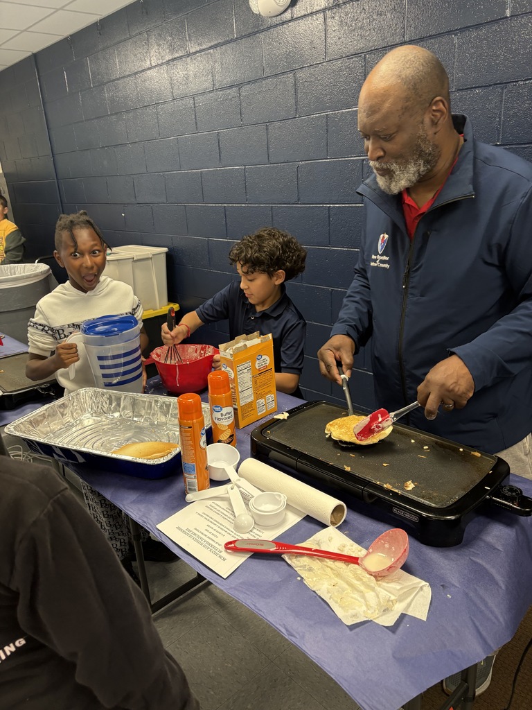A man cooks on a flat top griddle while two children observe. A table holds various cooking utensils and ingredients.