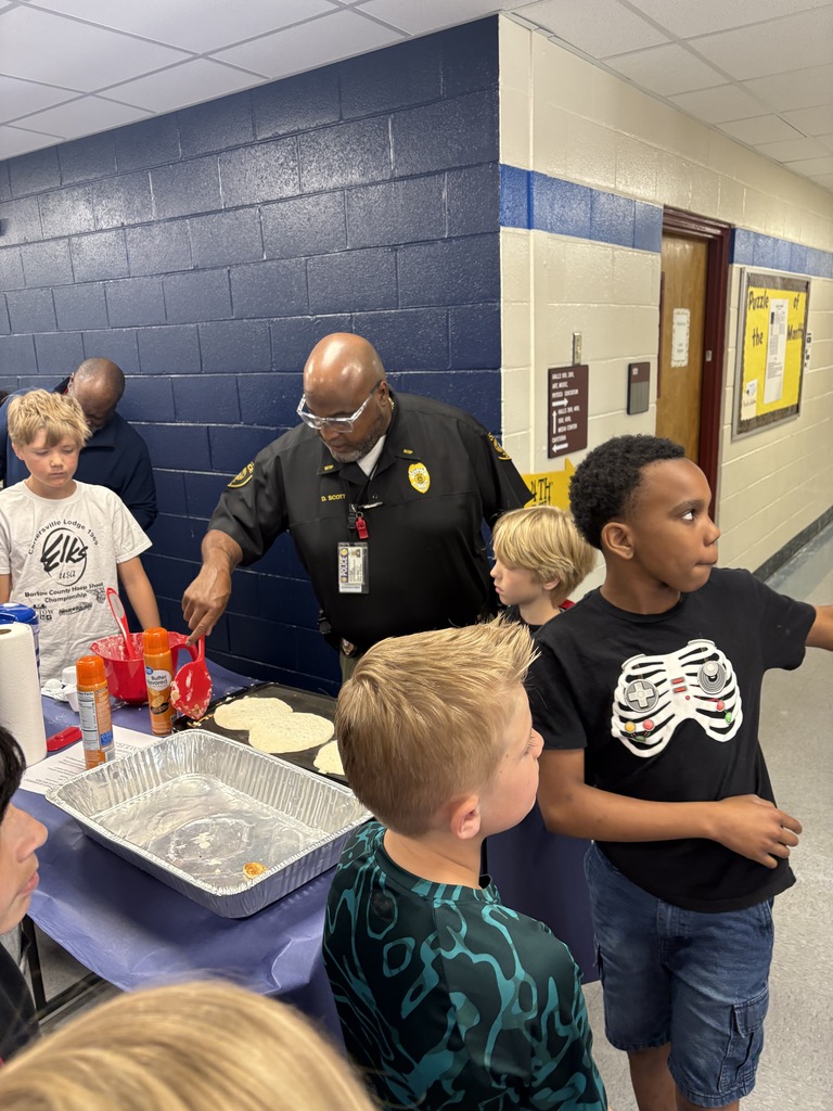 A uniformed officer stands at a table with children, a tray of food, and various containers.