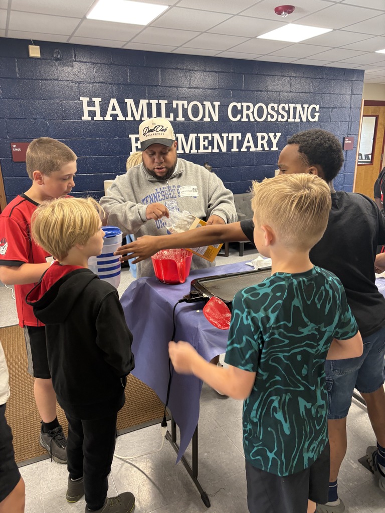 Several children surround a man standing at a table, reaching into a bowl. Behind them is a blue wall with the words "Hamilton Crossing Elementary."