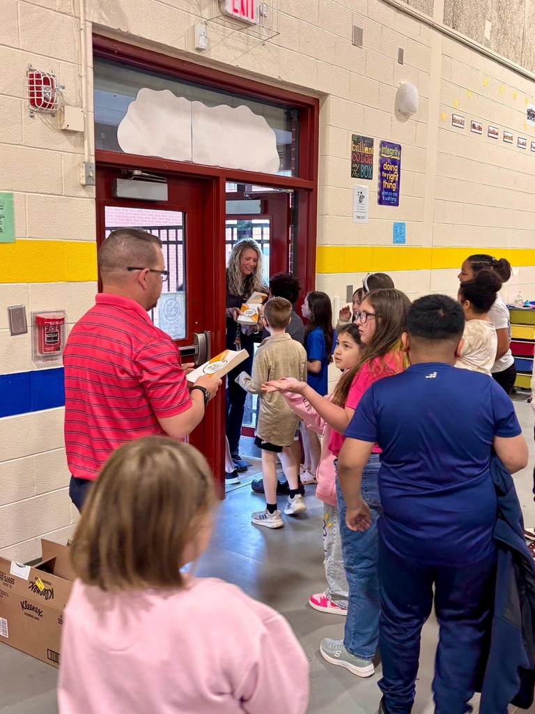 Adults and children gather around a man in a red shirt, holding food. Walls are yellow and white.