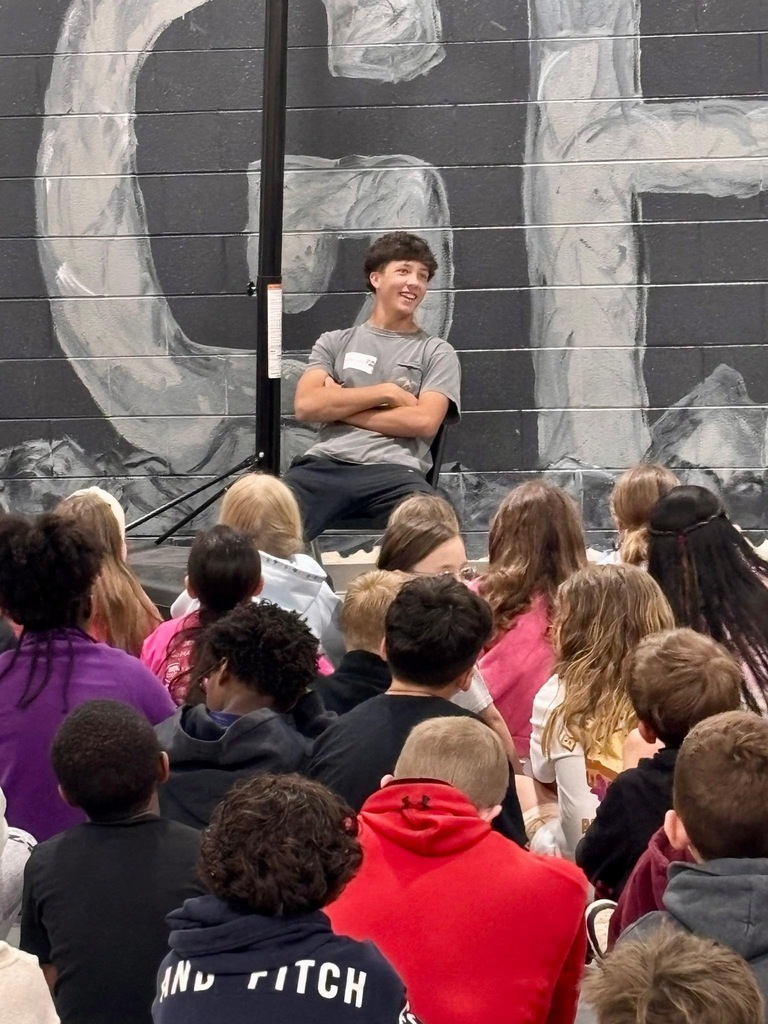 A person sits on a stage in front of a large audience, with a large "CH" on the wall behind.