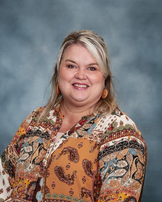 A woman with blonde hair smiles, wearing a colorful, paisley-patterned shirt. She stands against a blue background.