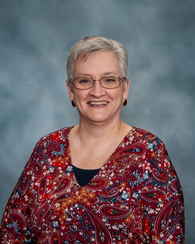 A smiling woman with short white hair wearing glasses and a paisley top. She stands against a light blue background.