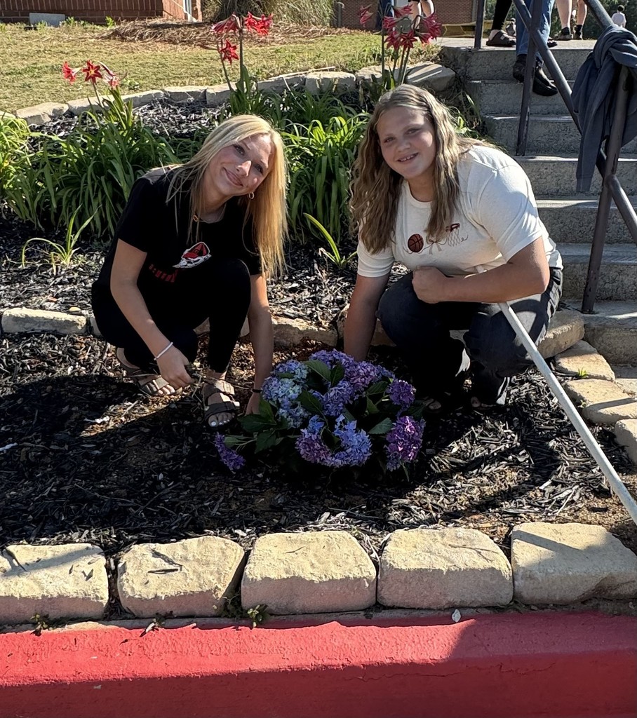 Two women planting flowers. One kneels on the ground, the other is seated. Behind them is a garden and stairs.