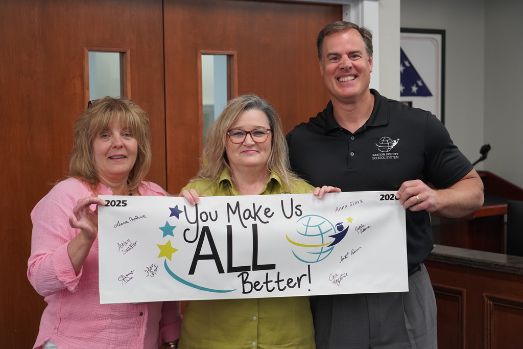 Three people pose with a white banner that reads, "You Make Us ALL Better!" The woman on the left wears a pink shirt.