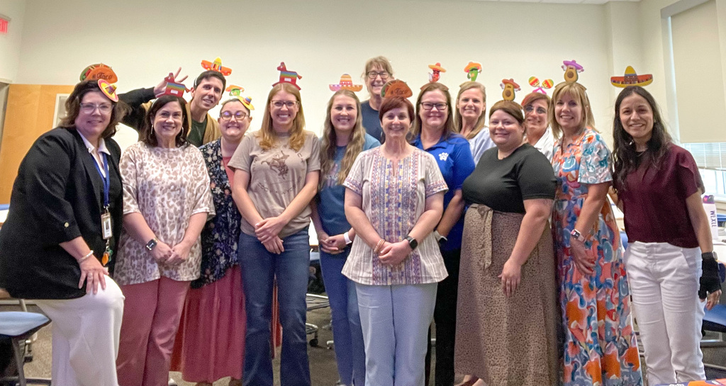A group of women wearing festive hats stands in a room with white walls, posing for a photo.
