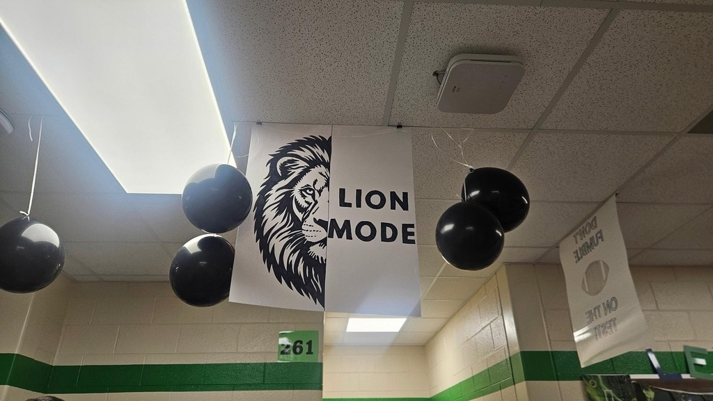 A classroom with white ceiling and green trim. A banner with "LION MODE" text and a lion logo hangs with black balloons.