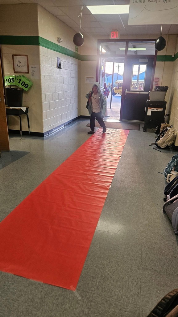 A hallway with a red carpet, a person in the middle, framed photo on the wall, open door, and a school bus visible outside.