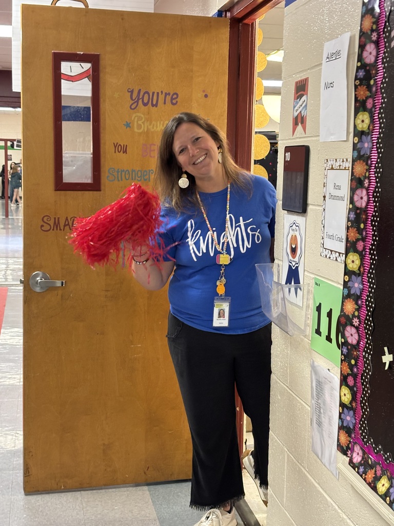A smiling adult woman in a blue shirt holds red pompoms. Behind her, a door and bulletin board with papers are visible.