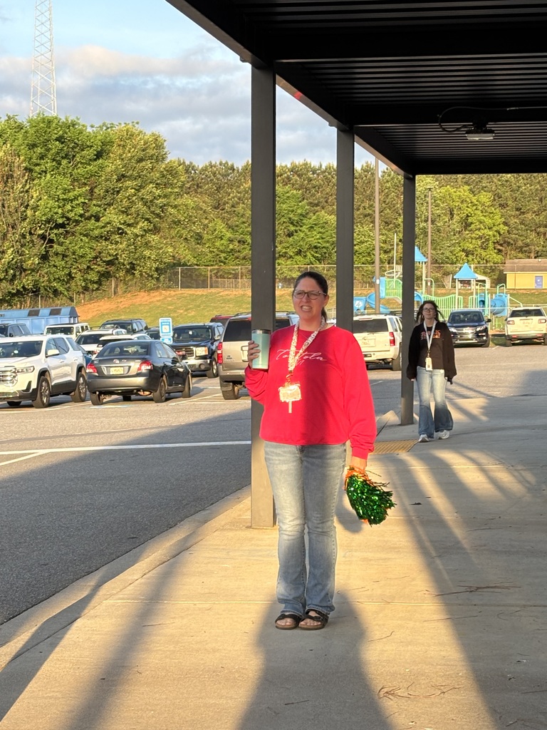 A woman stands in a school parking lot holding a bouquet. She wears a red sweatshirt and jeans. A person walks behind her, and many cars are parked.