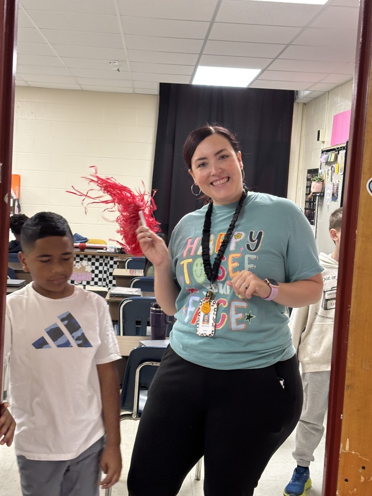 A woman holds red streamers and smiles at a boy in a classroom with chairs and a black curtain.