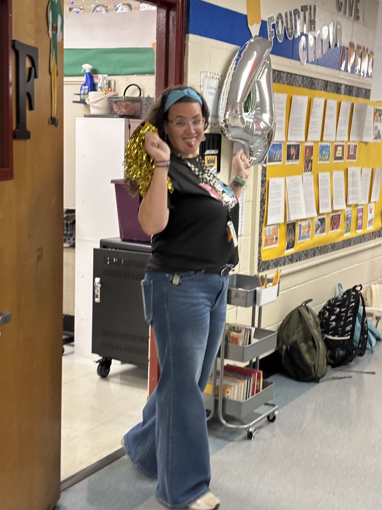 Woman with balloon and pom-poms stands in classroom hallway, facing camera. Behind her is a bulletin board and a cart.