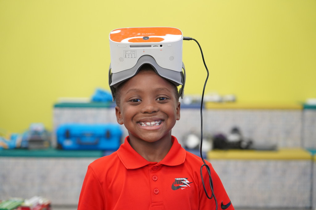 A smiling boy with a VR headset atop his head stands in front of a yellow wall.