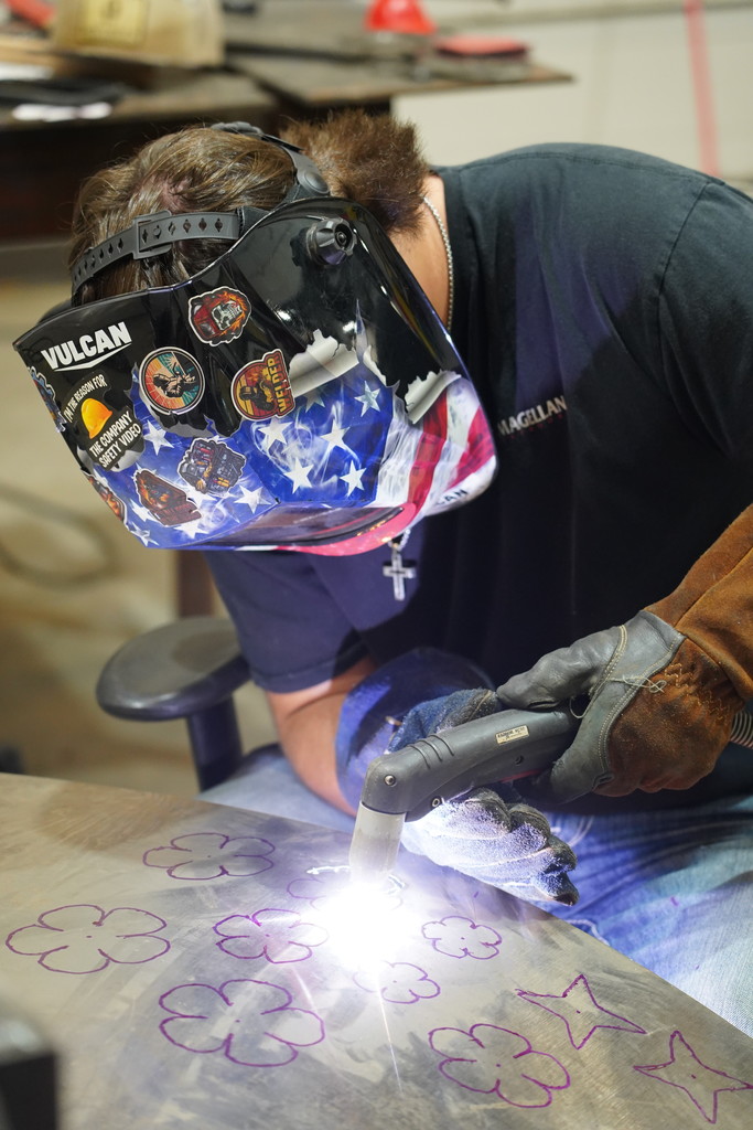 A person in a welding mask, gloves, and a shirt works on a metal sheet with a torch.