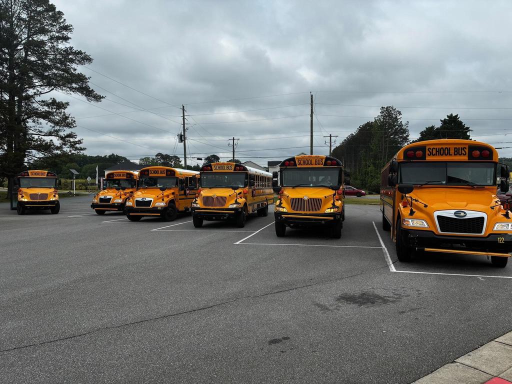 Multiple yellow school buses parked in a parking lot, under a cloudy sky with trees in the background.