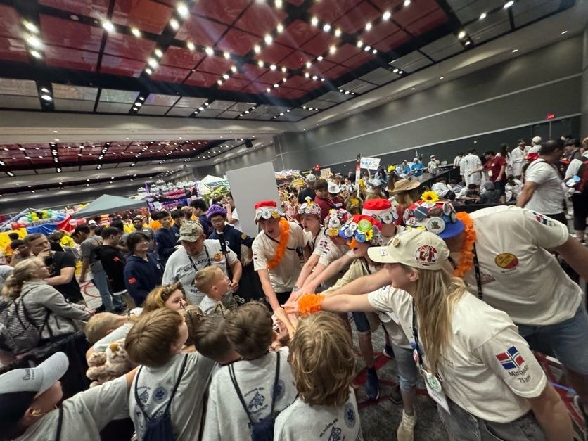Large indoor hall with people wearing white shirts and various hats, some touching hands. Background shows a high ceiling with lights.