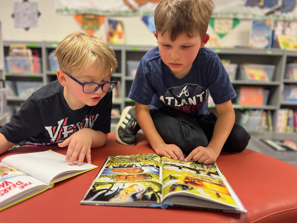 Two students seated in a school library reading books, with shelves of books surrounding them.