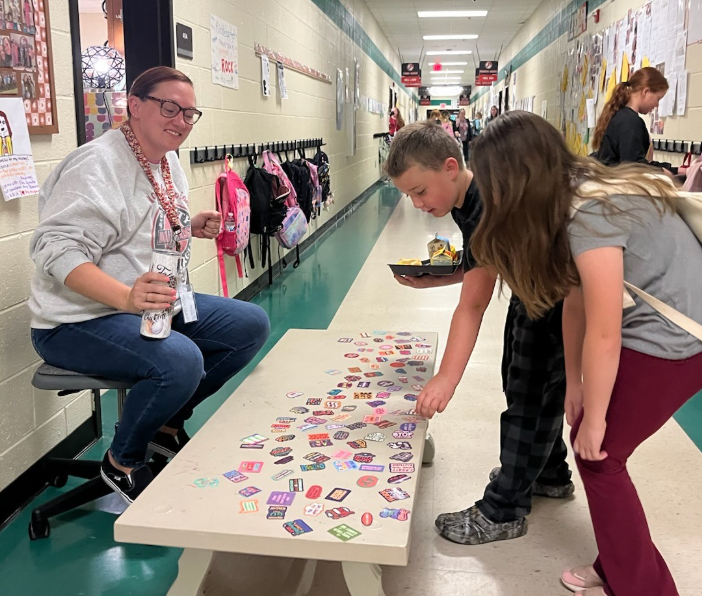 Two students standing beside a low table in a school hallway, selecting from a collection of small colorful stickers spread across the tabletop. Each student holds a cafeteria tray. An adult sits on a stool nearby holding a water bottle. Backpacks hang along the wall behind them, and additional people are visible further down the hallway.