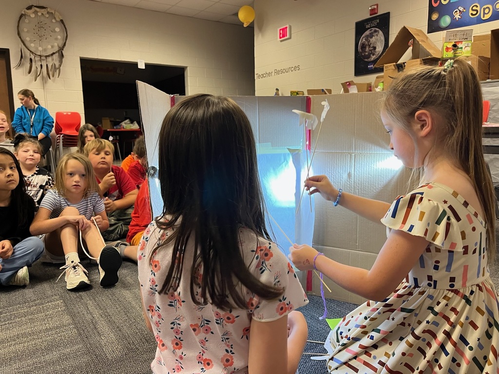 Students seated in a school library watch a puppet show performed behind a cardboard screen. Two students use flashlights to project light onto the screen while moving hand‑made puppets on sticks to create shadow images. Additional students sit on the floor in front of the stage area observing the performance. Bookshelves, posters, and classroom materials are visible in the background.