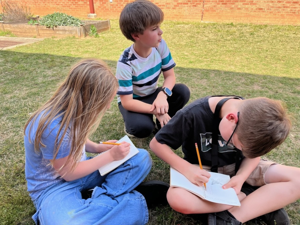 Three students sitting on grass outside near a brick school building, each holding a notebook and pencil. Two students are writing or sketching in their notebooks while another sits nearby. A raised garden bed and grassy area are visible in the background.