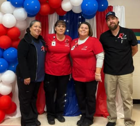 Four food service staff members stand together in front of a red, white, and blue balloon arch display indoors. Three individuals wear matching red shirts with name badges, and one wears a black shirt and cap. A table covered with a coordinating cloth is partially visible behind them.