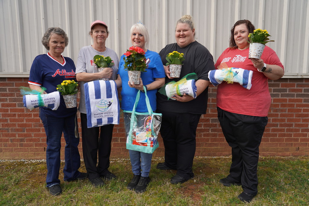 Six women stand in front of a brick wall holding potted plants and towels. They wear different colored shirts and pants.