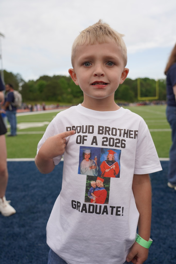 A boy wearing a white T-shirt with "Proud Brother of a 2026 Graduate" is pointing at the camera.