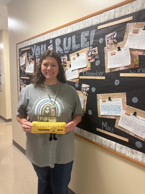 Person standing by a bulletin board holding a yellow rectangle with text. Board has pinned papers.