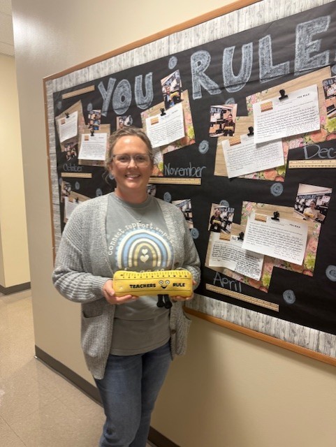 A woman stands next to a bulletin board holding a yellow object. Behind her is a bulletin board with several papers pinned to it.