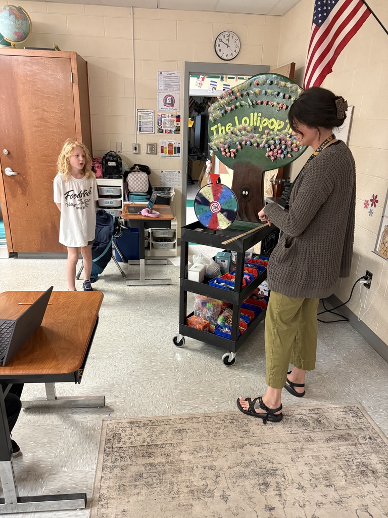 A teacher and a child are in a classroom, standing next to a cart with a spinner. The room has tables, a rug, and a flag on the wall.