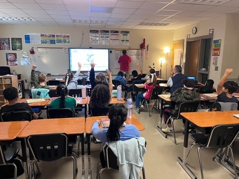 Classroom with desks and chairs. Students sitting, raising hands. Teacher stands by a projector screen.