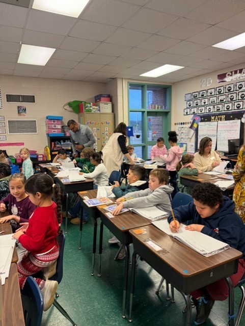 A classroom with students seated at desks, engaged in writing activities. A teacher is assisting students.