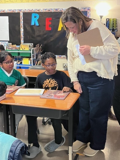 A woman stands in a classroom, looking at papers with two students sitting at a desk.