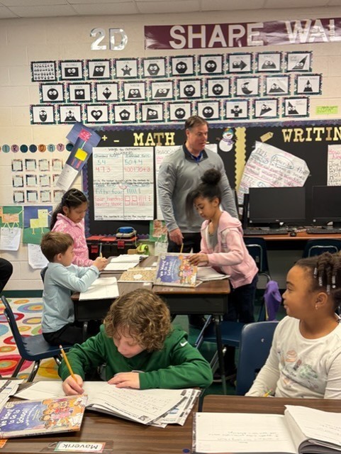 Children seated at desks in a classroom, working on writing. One child leans over a desk, writing.